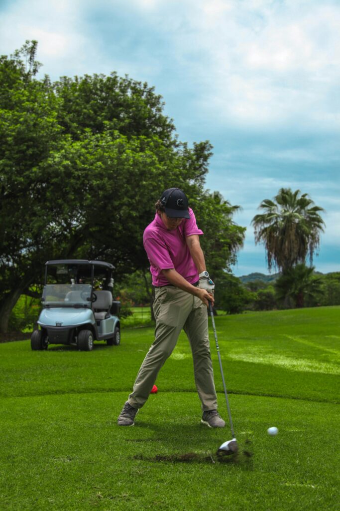 Male golfer in action on a green course, focusing on swing with cart nearby.